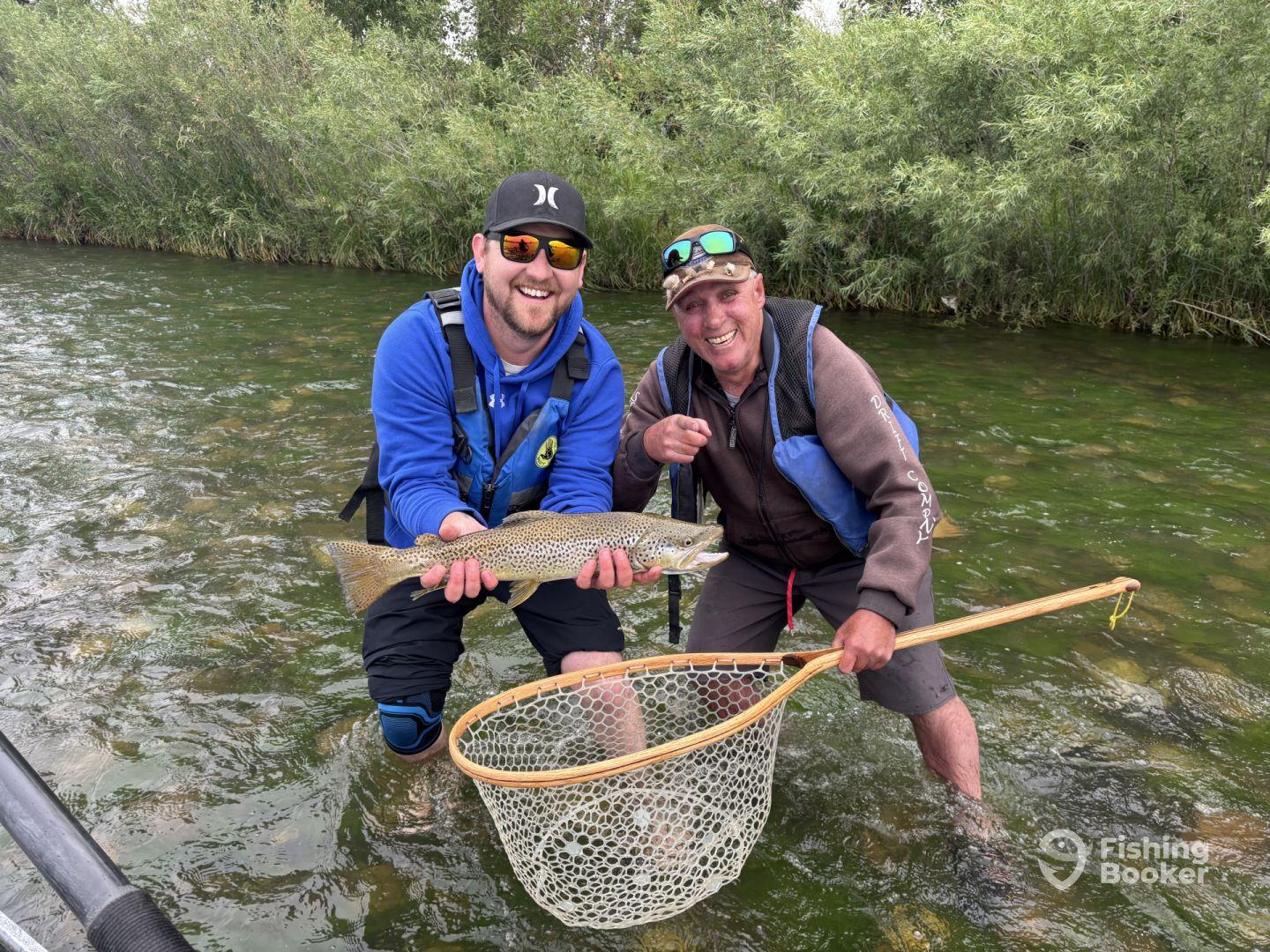 Two anglers proudly displaying a large Brown Trout while fishing in a scenic river environment.