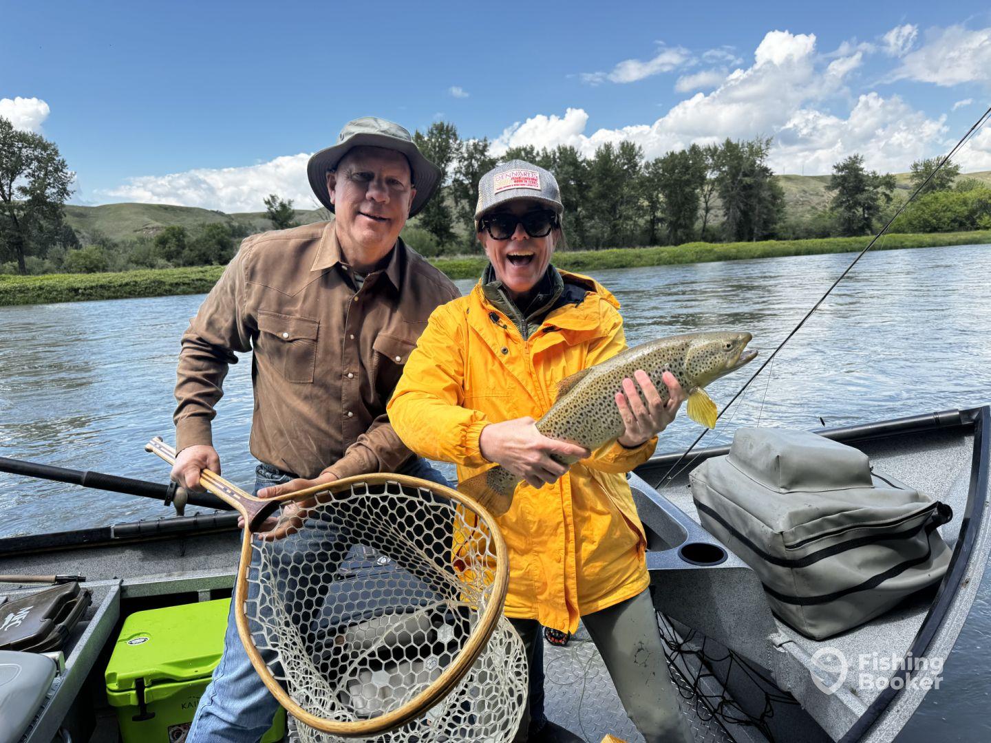 Two anglers celebrating a successful catch of a large Brown Trout while fishing on a scenic river.