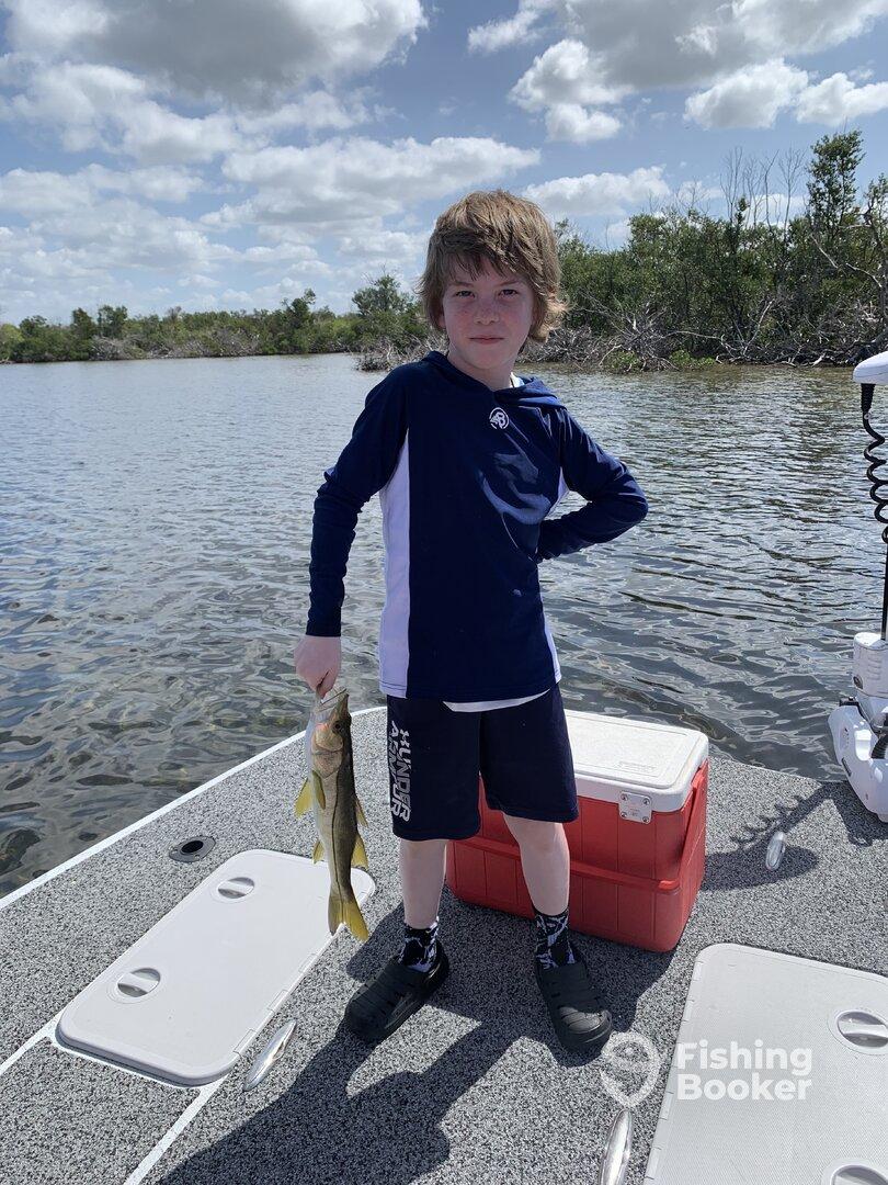 Young angler proudly holding a Snook while fishing on a boat in a scenic waterway.