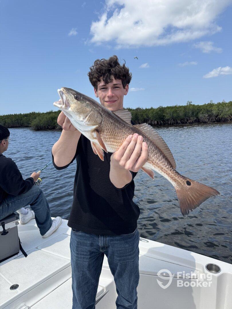 A young angler proudly displaying a Redfish while fishing on a boat in a scenic waterway.