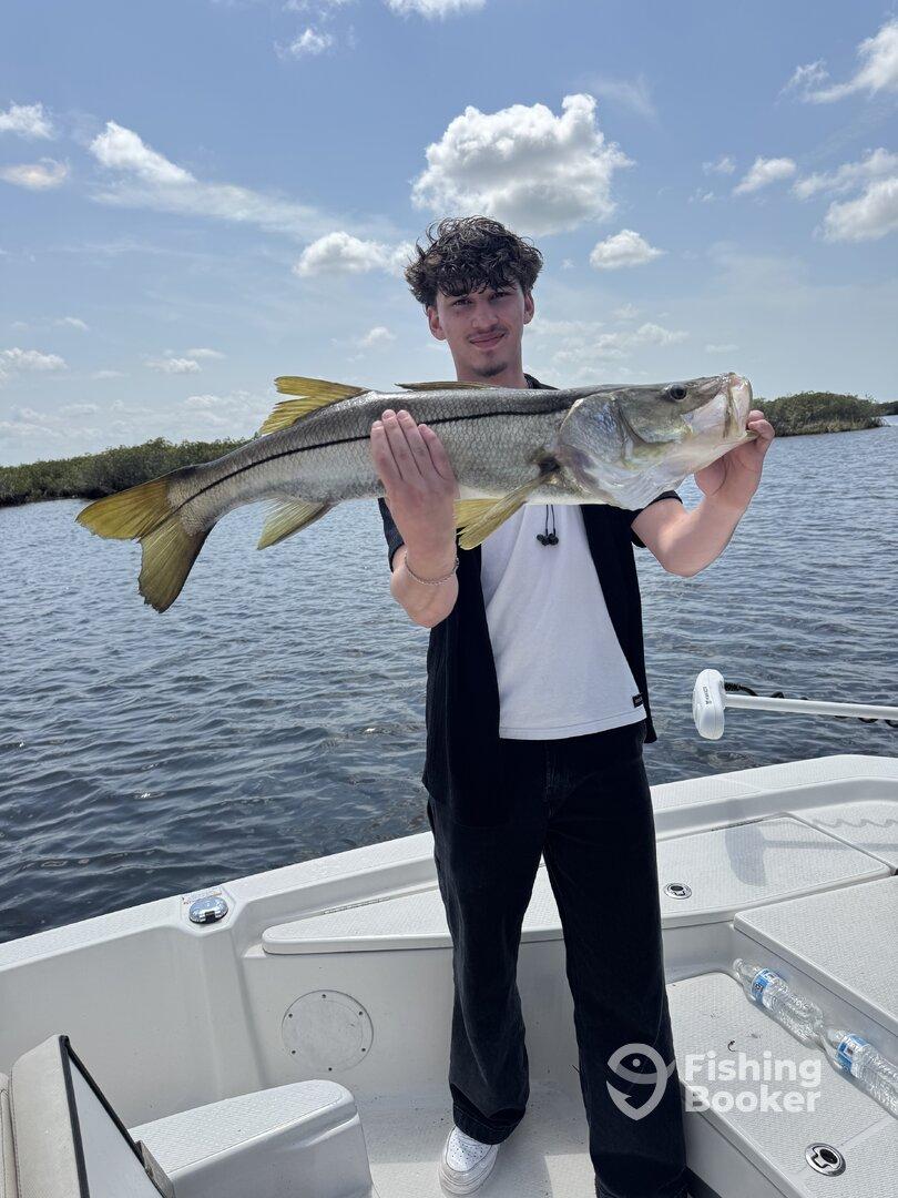 A young angler proudly displaying a large Snook while fishing on a boat in a scenic waterway.