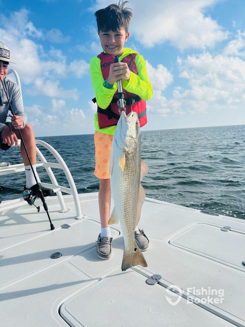 A young angler proudly displaying a large Redfish while fishing on a boat, showcasing a sunny day on the water.