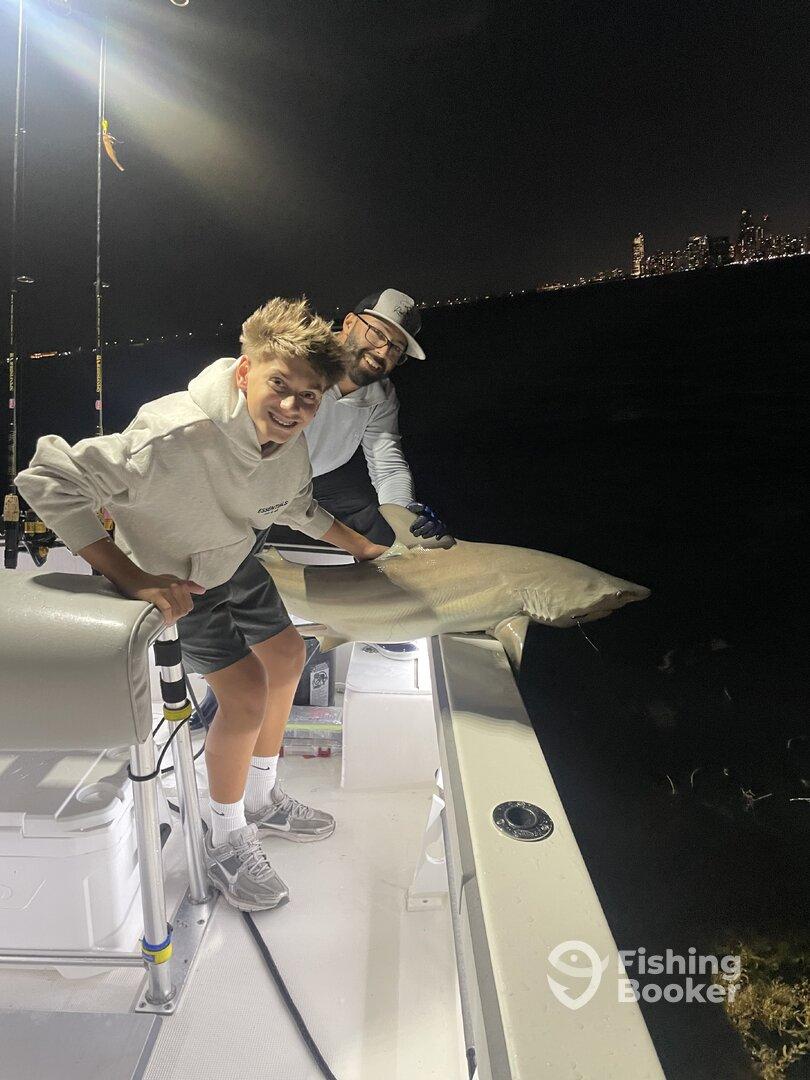 A young angler and an adult proudly displaying a caught shark while fishing at night, with a city skyline in the background.