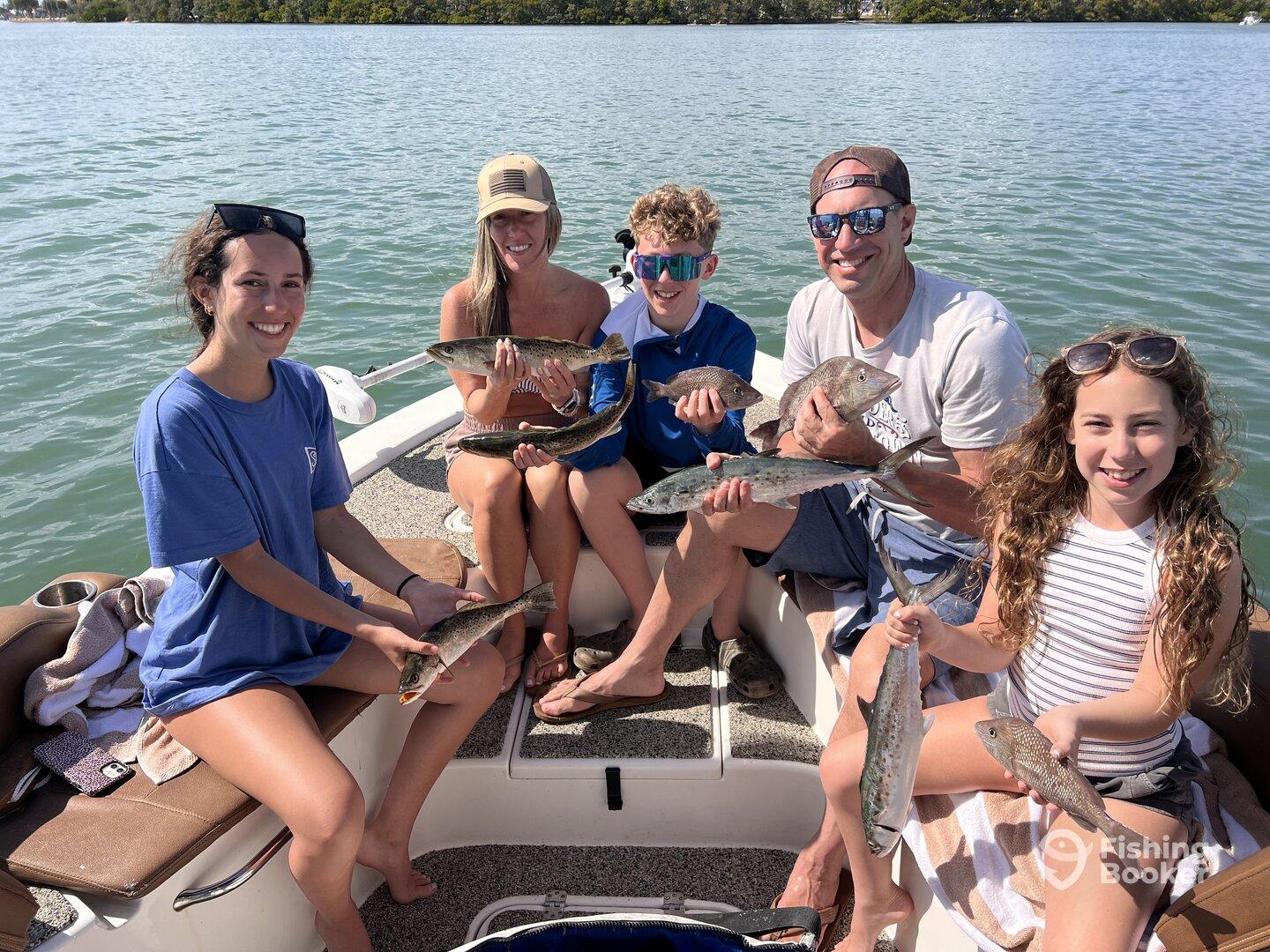A family enjoying a successful fishing trip, proudly displaying their catch of various fish species while on a boat in a scenic waterway.