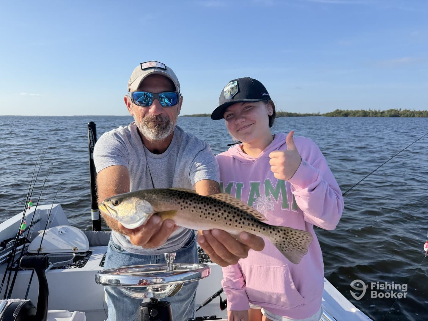 A young angler and an adult proudly displaying a Speckled Trout while fishing on a boat in a scenic waterway.
