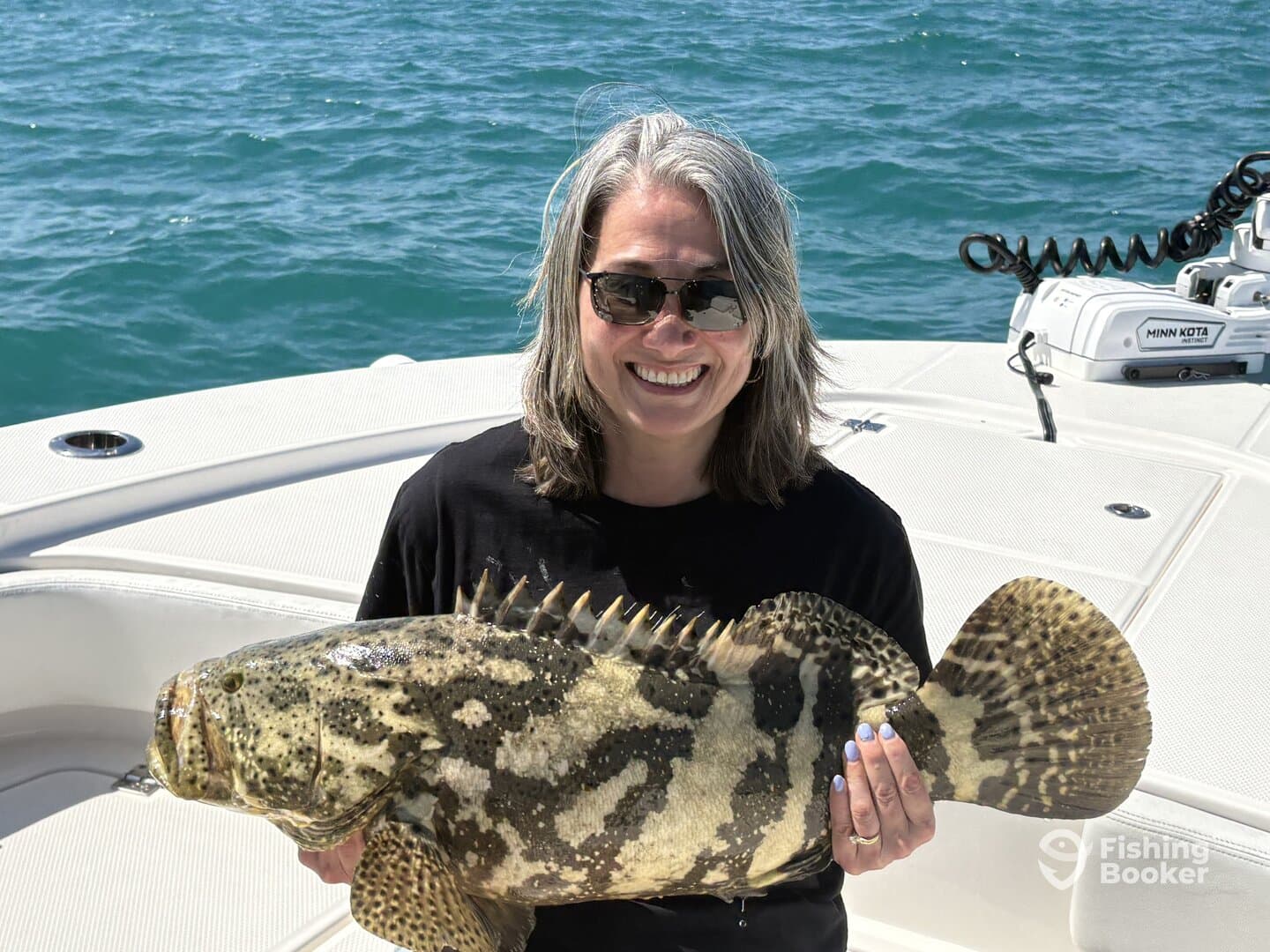 A smiling angler proudly displaying a large Grouper while on a fishing boat, showcasing a successful day on the water.