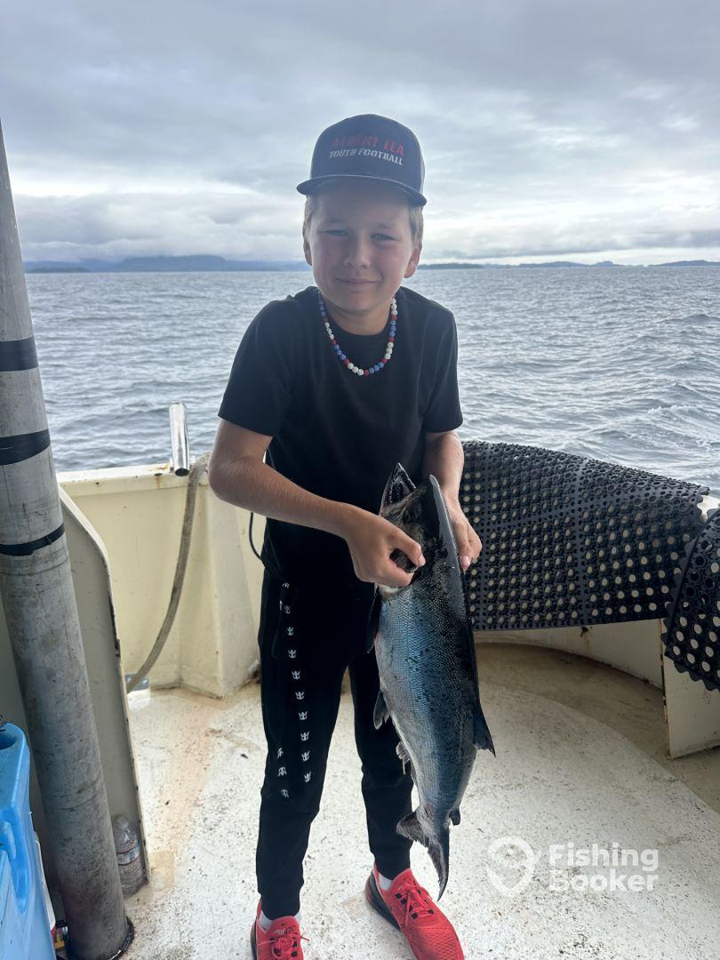 A young angler proudly displaying a freshly caught Salmon aboard a fishing boat, surrounded by a scenic ocean backdrop.