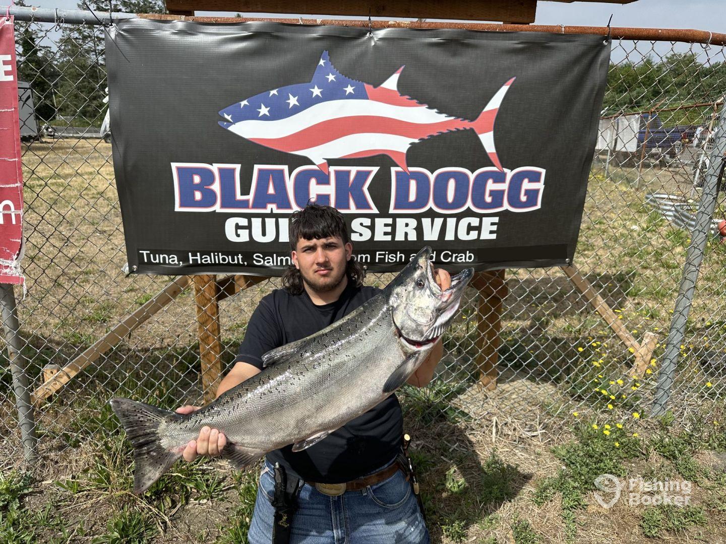 Angler proudly displaying a large Salmon in front of the Black Dogg Guide Service sign, showcasing a successful fishing trip.