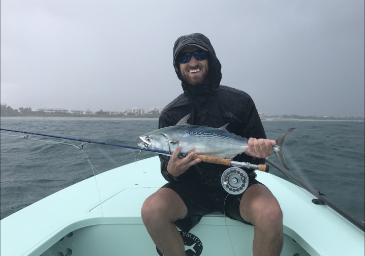  en smilende fisker i en regnjakke og shorts holder en falsk albacore og en fluefiskestang på en båt.