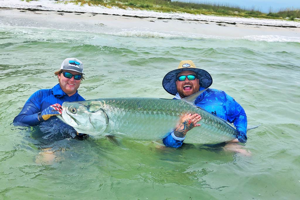 Two smiling men in caps and sunglasses standing in water with a Tarpon in their hands
