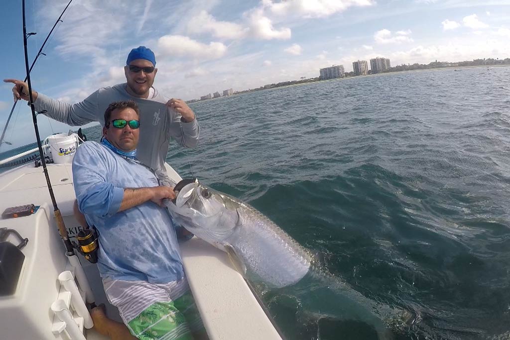 Two fishermen standing on a boat, holding onto a big Tarpon still in the water, with Florida shore in the background