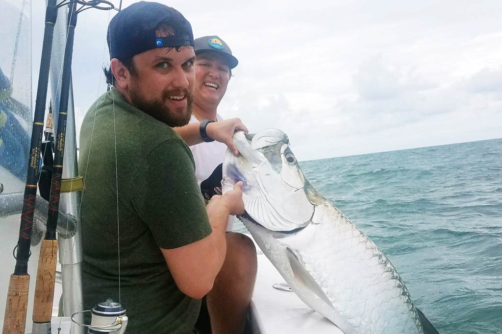 A fisherman and his friend looking at a camera while holding a Tarpon half-raised from the water