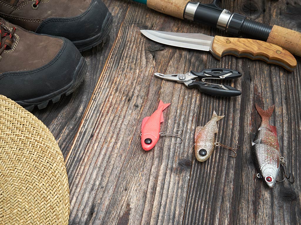 A selection of fishing equipment: a knife, pliers, lures, boots, fishing rod, and hat, all laid out on a wooden floor.