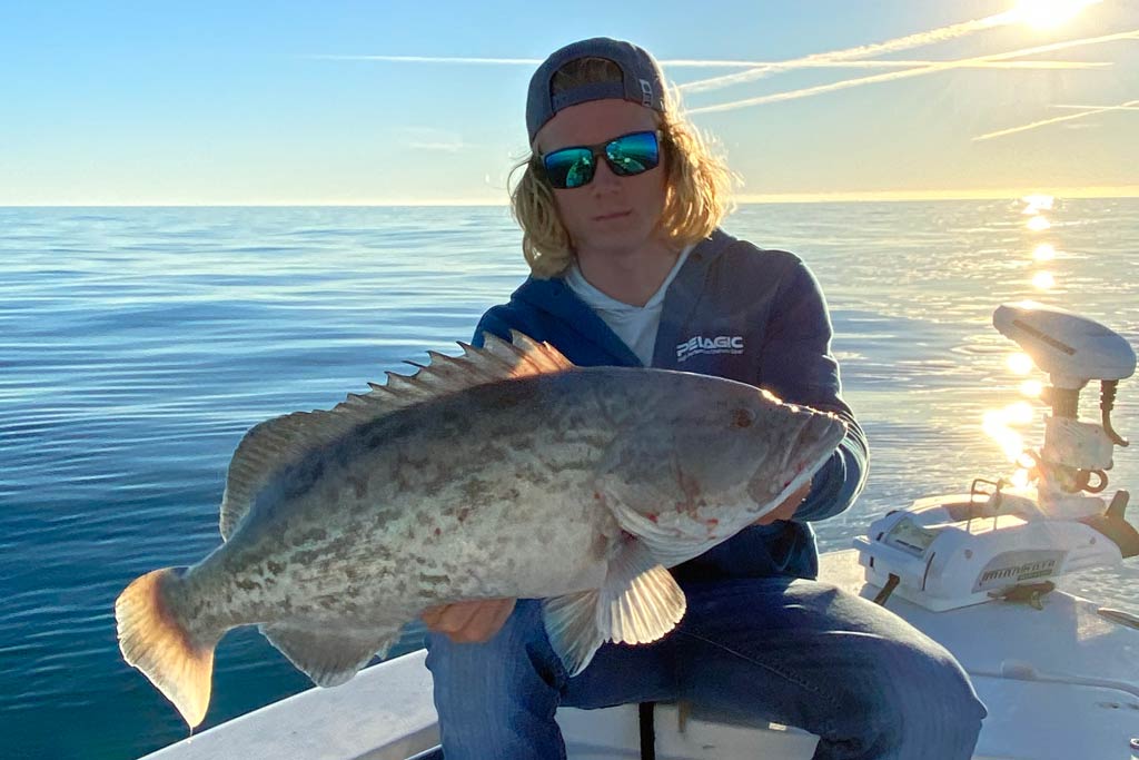 A young charter guide sitting on his fishing boat in a reversed cap and sunglasses, holding a Black Grouper he caught, with calm waters, blue skies, and the sun behind him.