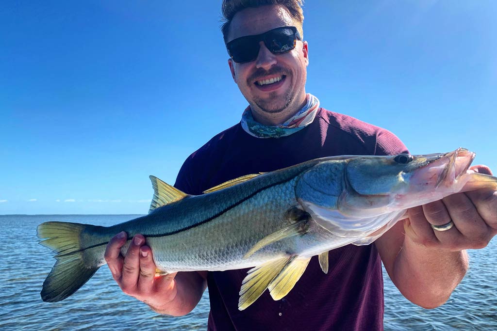 A smiling angler in sunglasses holding a Snook he caught, with blue skies in the background