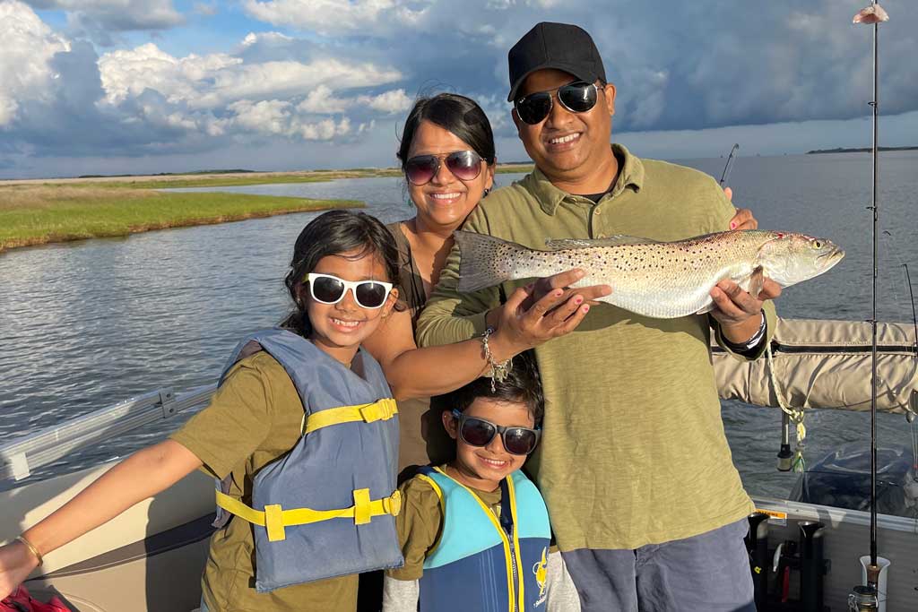 A smiling family in sunglasses standing on a fishing boar, father holding a Spotted Seatrout
