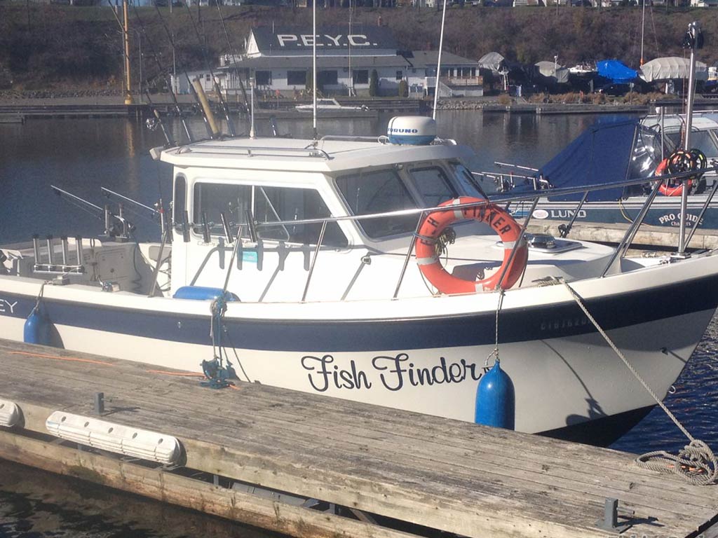 A photo of a pilothouse boat docked next to a wooden pier in the marina and surrounded by other charter fishing boats