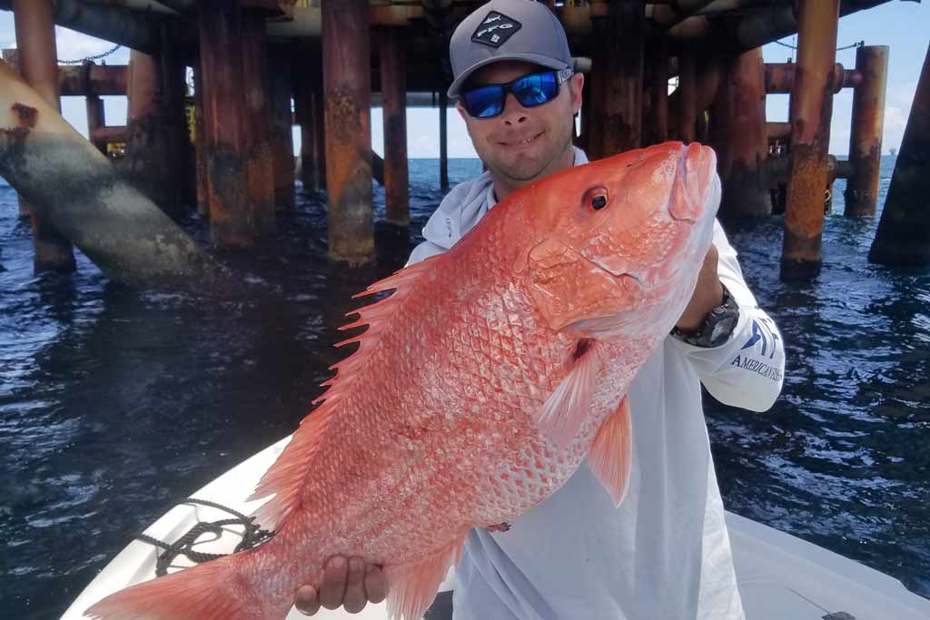A smiling angler standing on a boat, with an oil rig behind him, holding a big Red Snapper