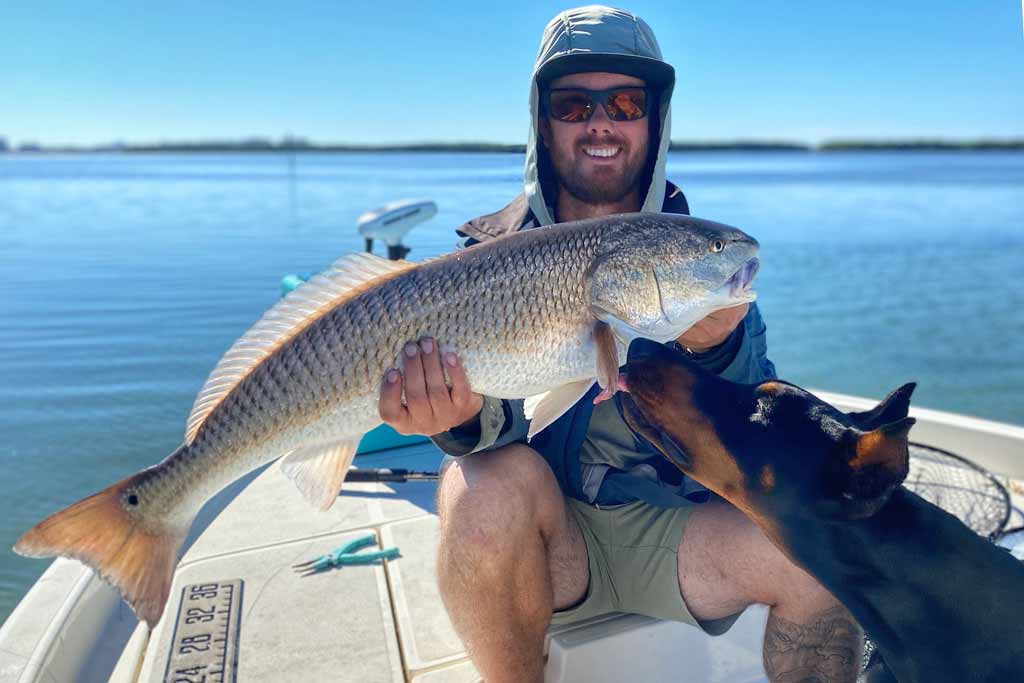 A smiling fisherman in a hat and sunglasses, sitting on a boat, holding a Redfish with a Doberman smelling the fish, with blue waters and skies in the background