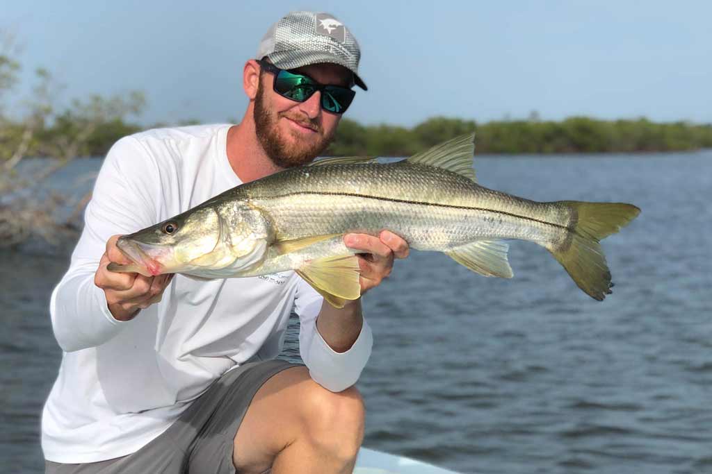 A smiling angler holding a large Snook while crouching on a boat with vegetation and opent waters in the background