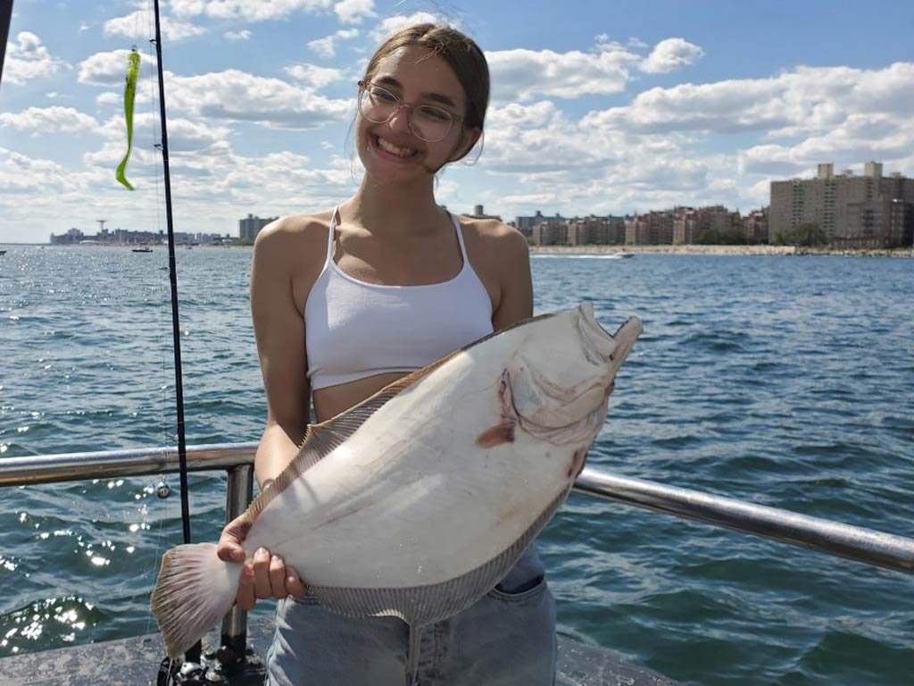 A photo of a female angler holding Fluke with both hands and smiling while standing on a charter fishing boat and posing against the skyline of the city in the background
