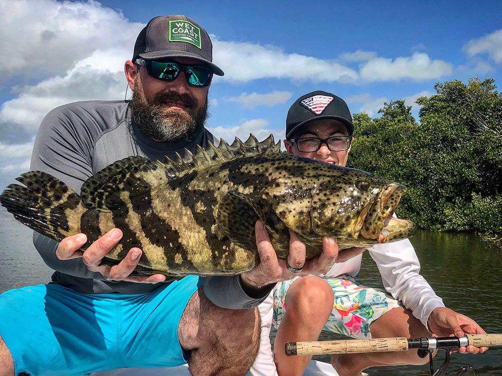 A man and a young child, sitting on a shallow-water fishing boat, posing with their catch of a Grouper with trees visible along the shore next to them on a sunny day