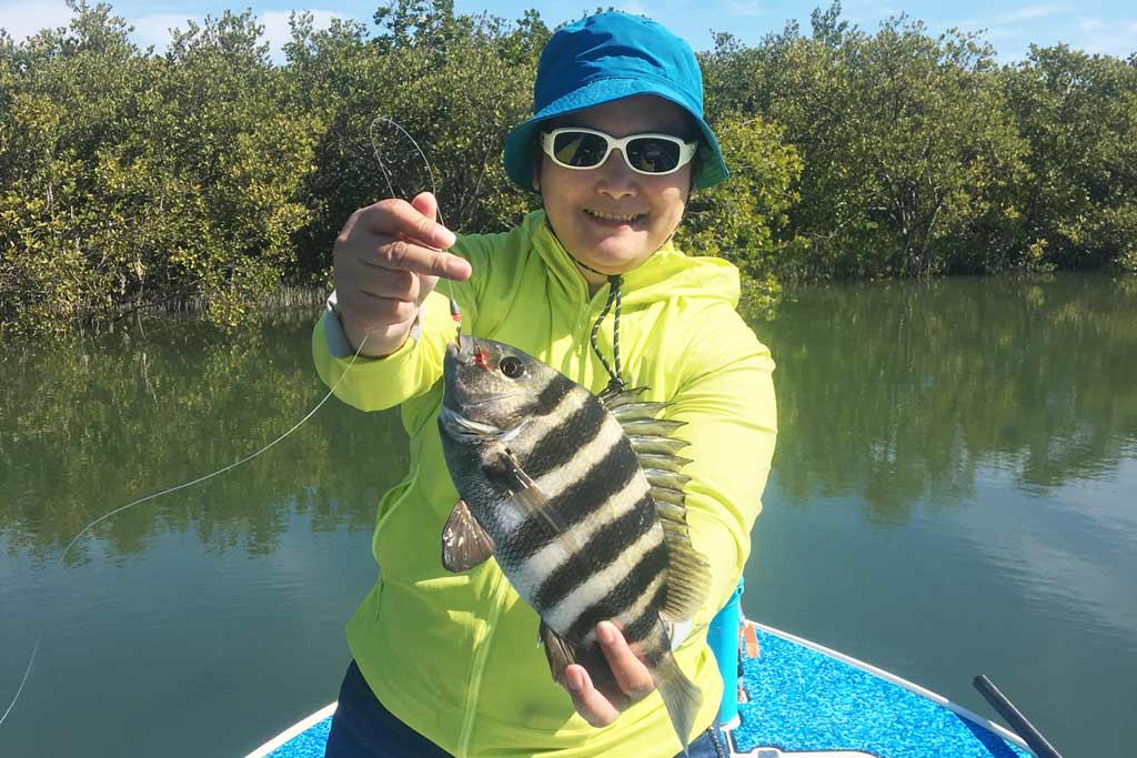 A smiling woman in a cap, sunglasses, and a fluorescent jacket holding a Sheepshead while standing on a fishing boat, with vegetation and water in the background