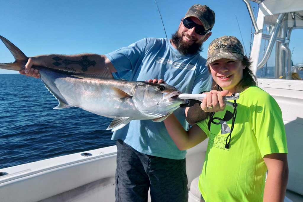 A charter guide in a cap and sunglasses standing next to a you angler in a cap and a fluorescent shirt, holding a big Amberjack while standing on a boat