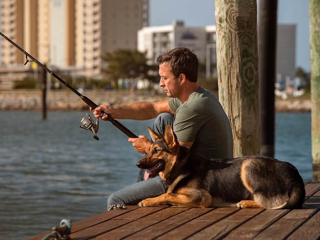 A photo of an angler sitting on a wooden dock, holding a fishing rod, and fishing while his dog is sitting next to him and looking at the water