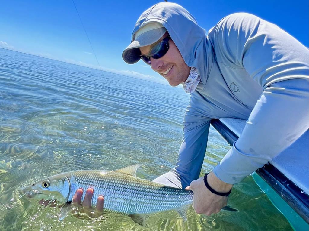 A side view of a smiling angler holding Bonefish with both hands and releasing it back to the water
