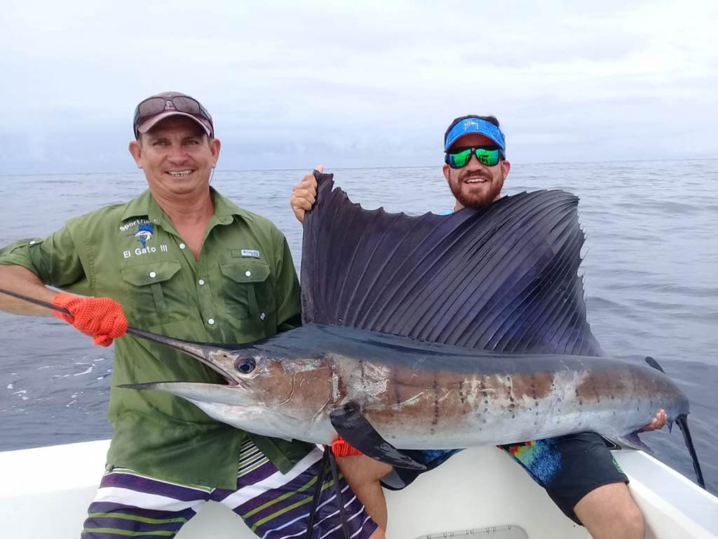 A photo of two anglers sitting on a charter boat and holding big Sailfish they caught while fishing offshore