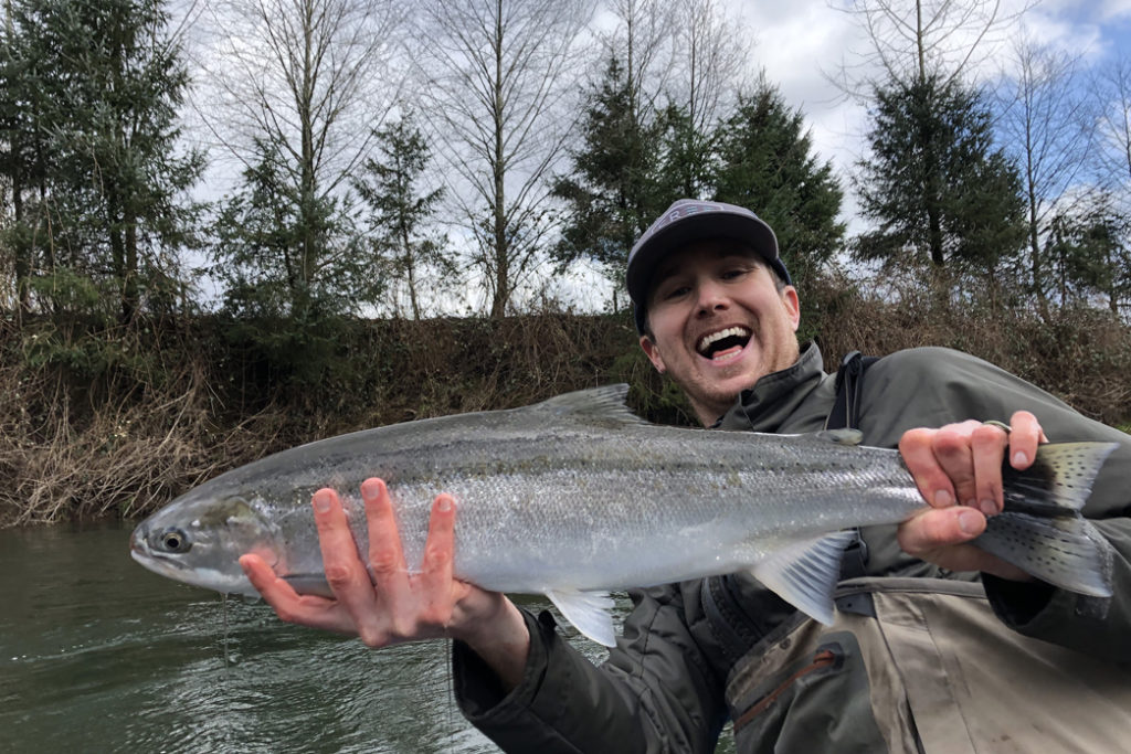 A man in a hat smiling and leaning back while holding a sizeable Steelhead he caught fishing in Oregon, with the river and shoreline foliage behind him.