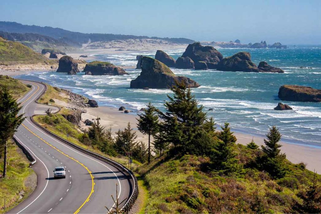 A view of Cannon Beach in Oregon, with a shoreline road to the left of the photo and the ocean to the right, with several rock formations sticking out of the water.