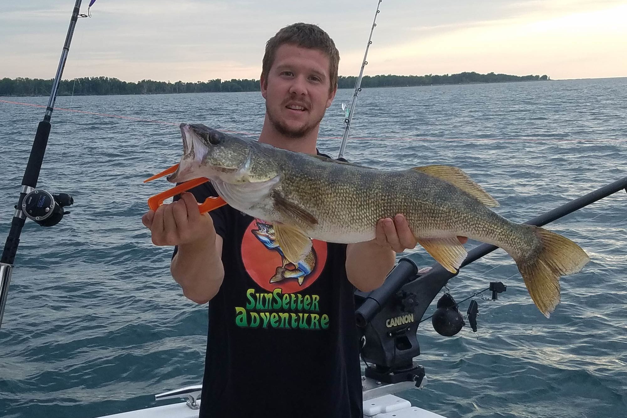 A young man standing in the corner of a boat, holding a big Walleye, with water in the background