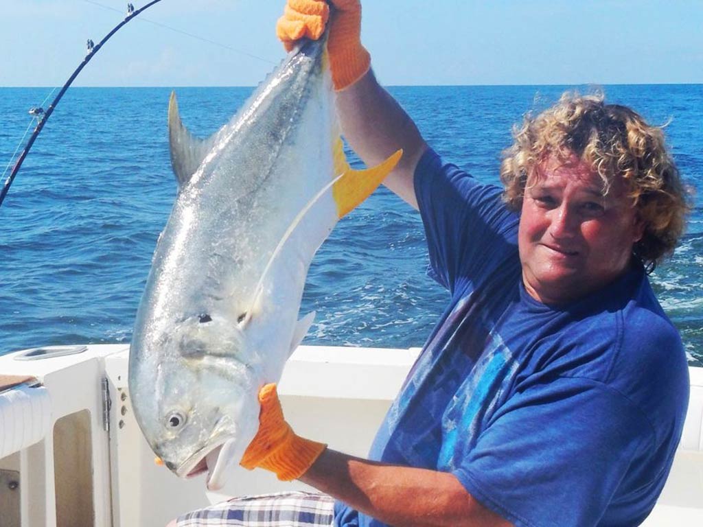 An angler with curly blonde hair holding a Jack Crevalle upside down, while standing on a fishing charter with the open waters behind him