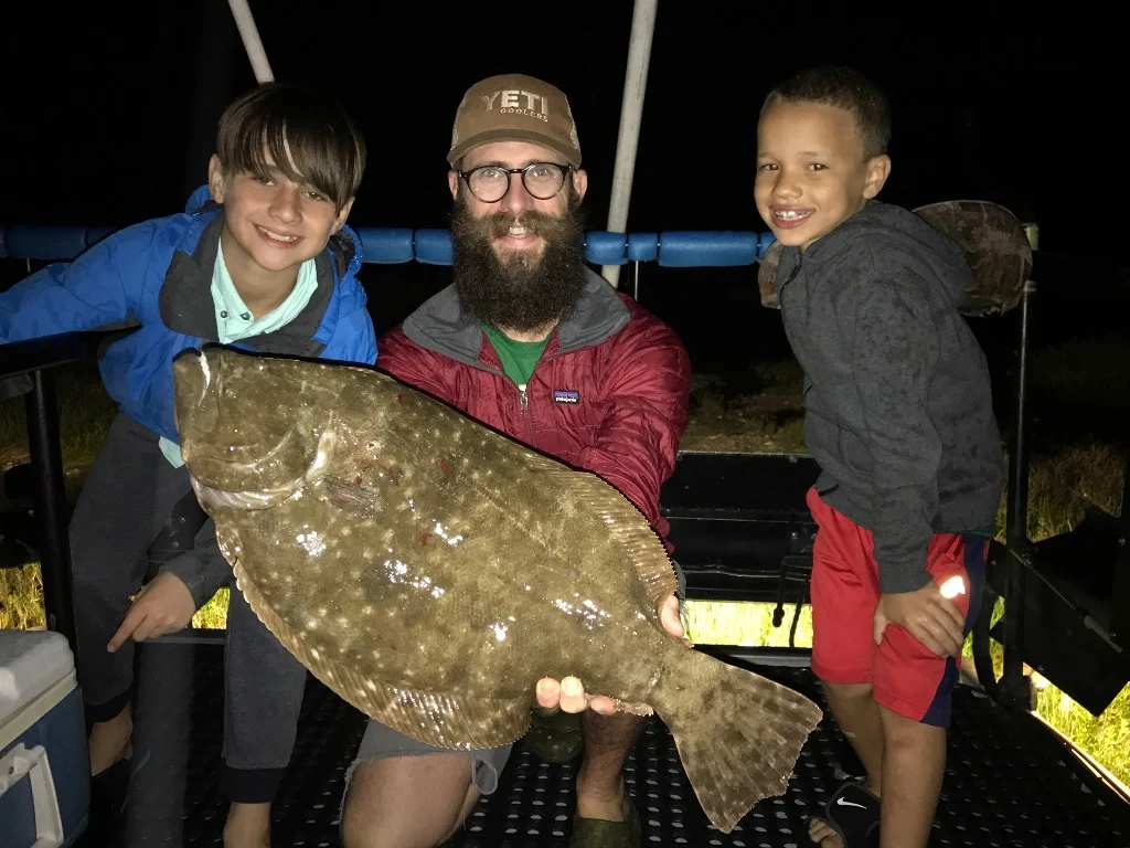 A happy bearded angler with two kids either side of him on a boat holding a Flounder caught by gigging at night