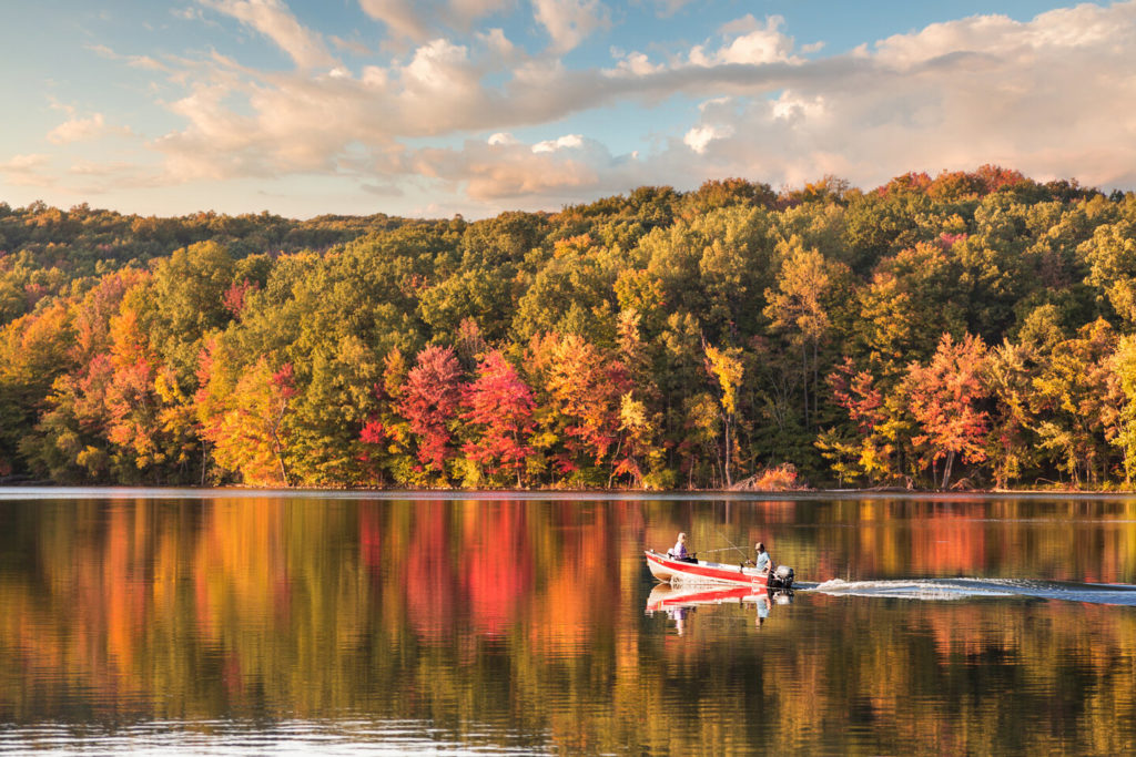 Two fishermen on a boat riding through a lake in Connecticut during the fall months with a forest in the background