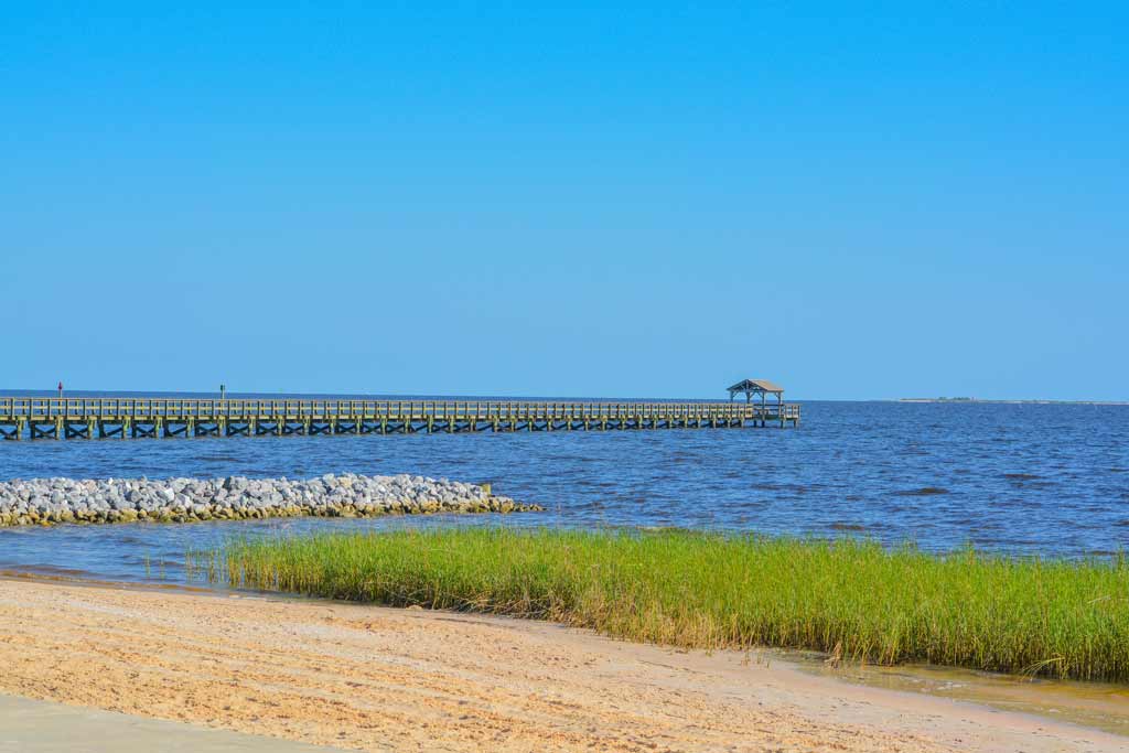 A long fishing pier in Biloxi in the distance, with water, a beach, and blue skies