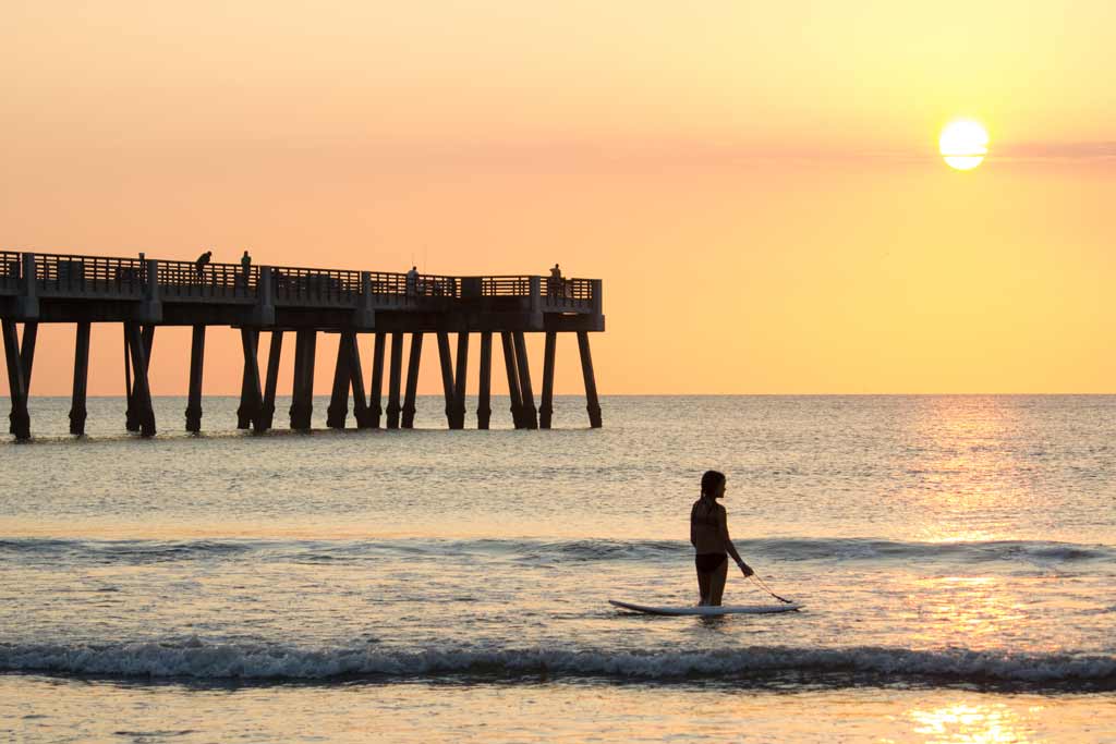 A woman with her paddleboard in the surf on a Jacksonville beach, with a pier in the background