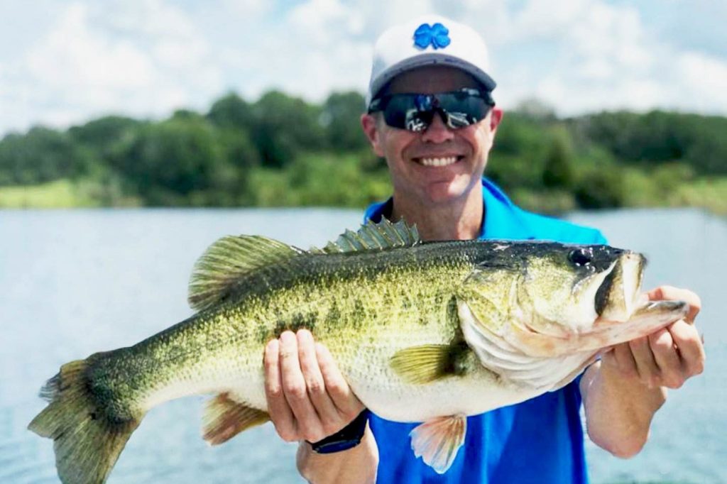 An image of a man wearing sunglasses holding a Largemouth Bass to the camera in Florida, with water overdue him and some foliage in the distance