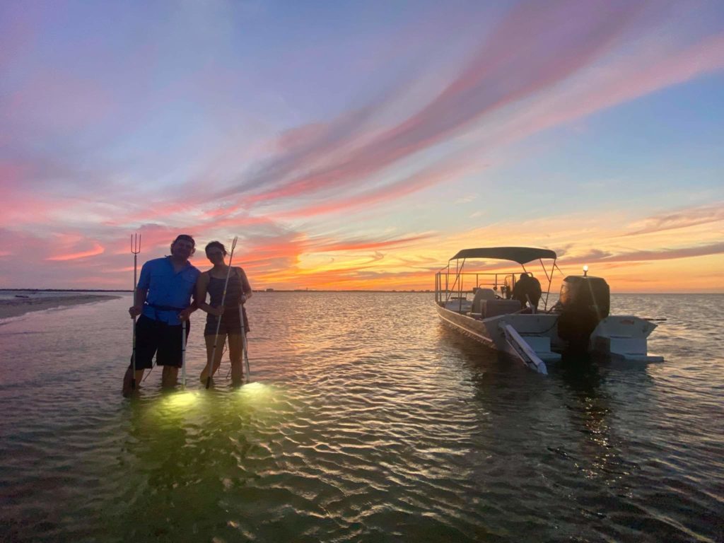 A male and female angler standing in the water by their boat with Flounder gigs and flashlights in their hands at sunset