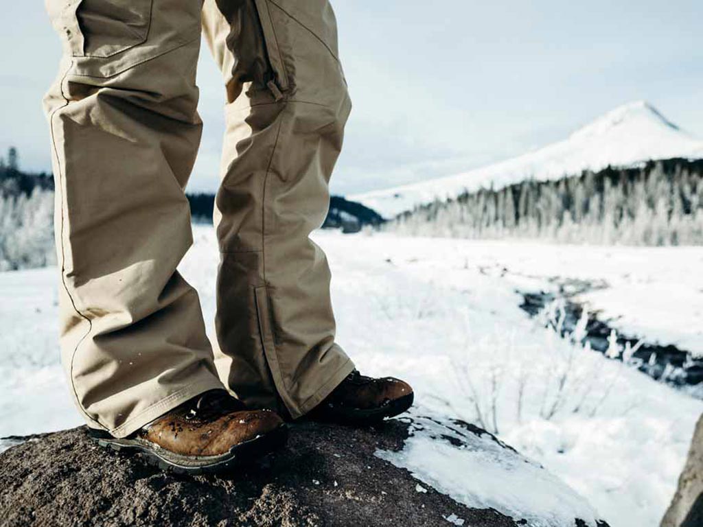 A closeup of man's waterproof pants while standing on a rock with snowy landscape in the background