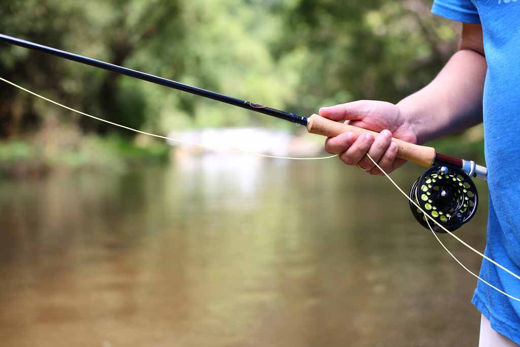 A close-up of an angler's hand holding a fly fishing rod with some slack, line, river in the background