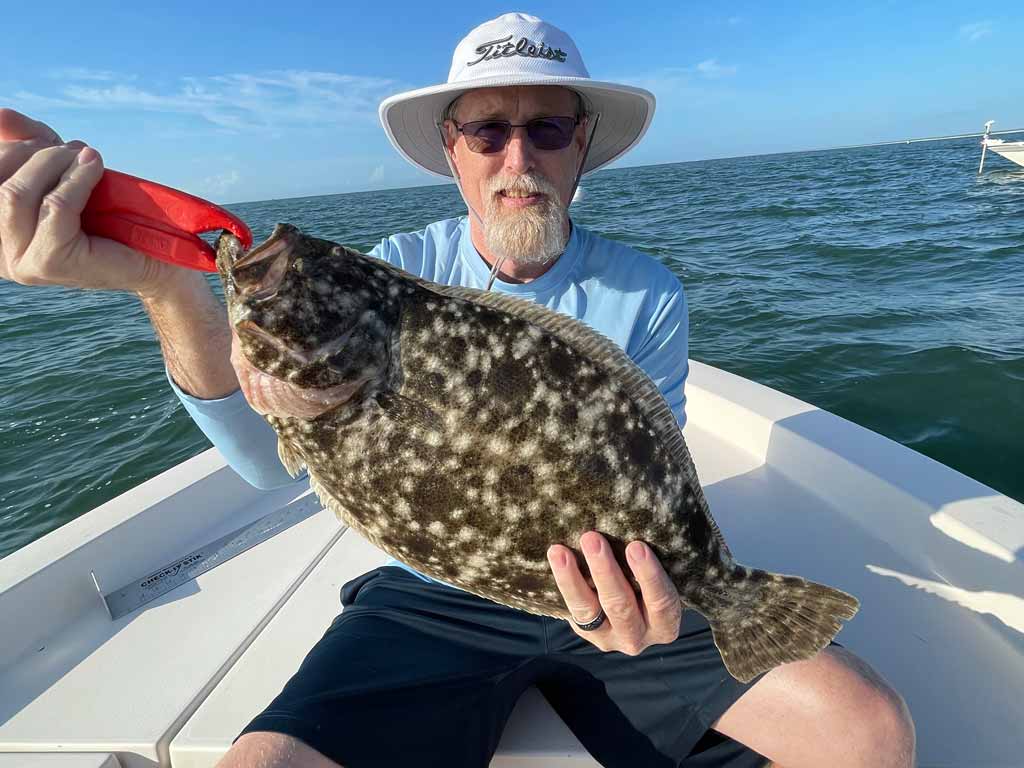 An older fisherman in a fishing hat and sunglasses, sitting on a boat, holding a big Flounder