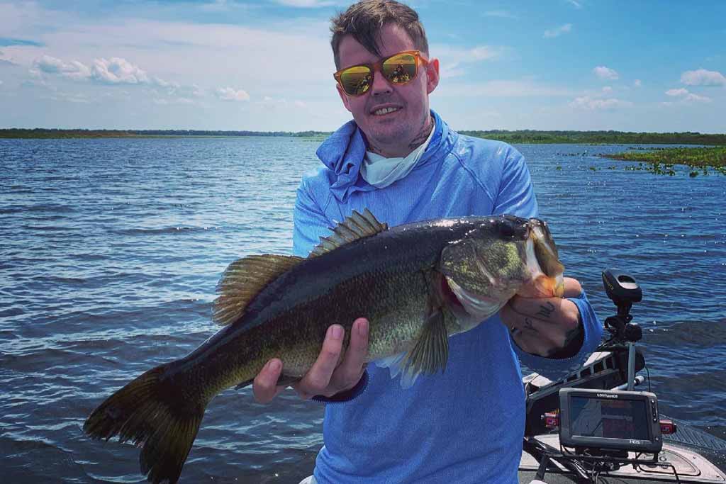 A man in sunglasses, standing on a boat, holding a big Largemouth Bass