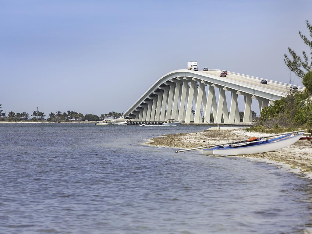 A side view of the Blind Pass Bridge leading out of Sanibel posing against the water and a tiny sandy beach featuring a small boat
