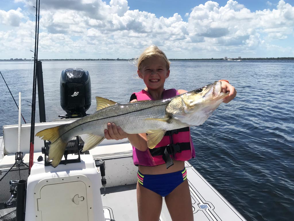 A photo of a young girl wearing a vest and standing on Sanibel charter fishing boat while holding a big Snook  