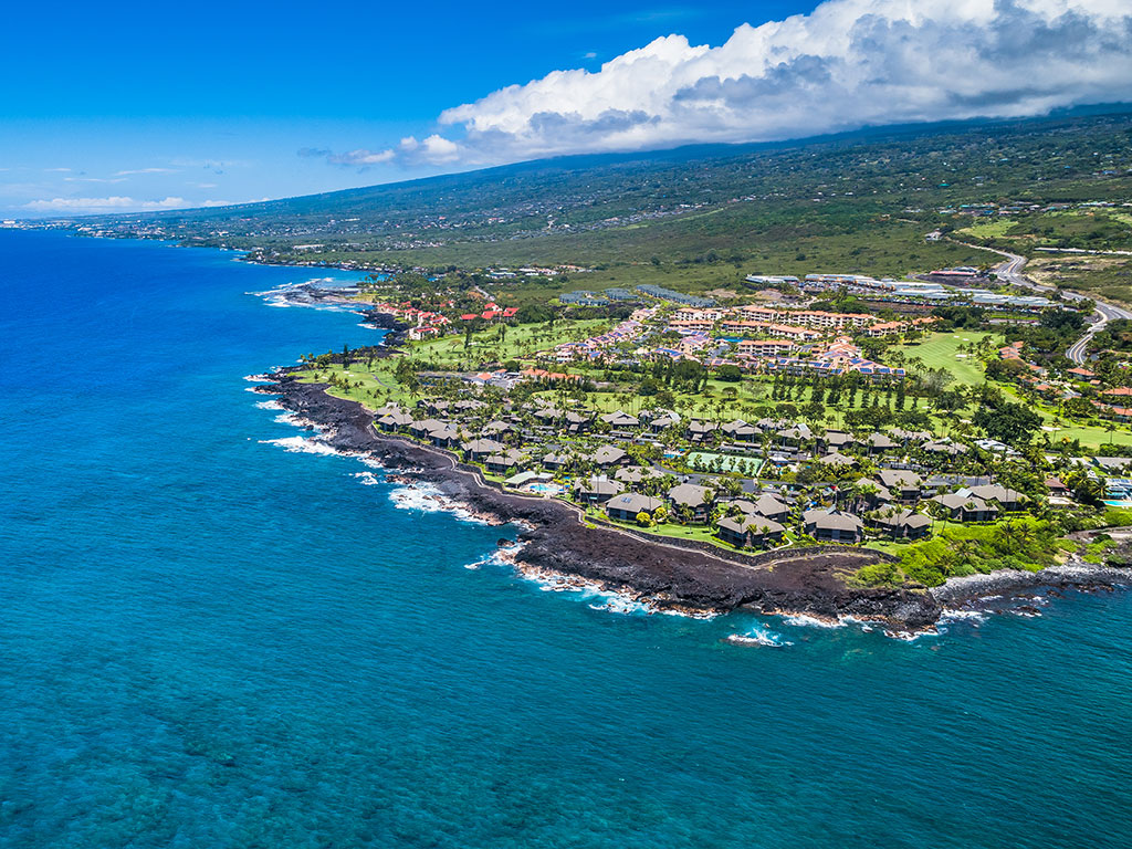 An aerial view of Kailua-Kona with the blue seas in the foreground and right and climbing land towards the mountain on the left