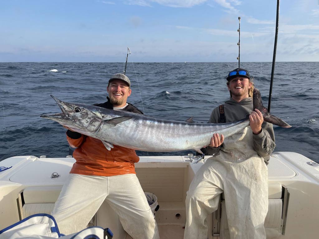 Two anglers on a boat, holding a huge Wahoo they reeled in while deep sea fishing in Outer Banks.