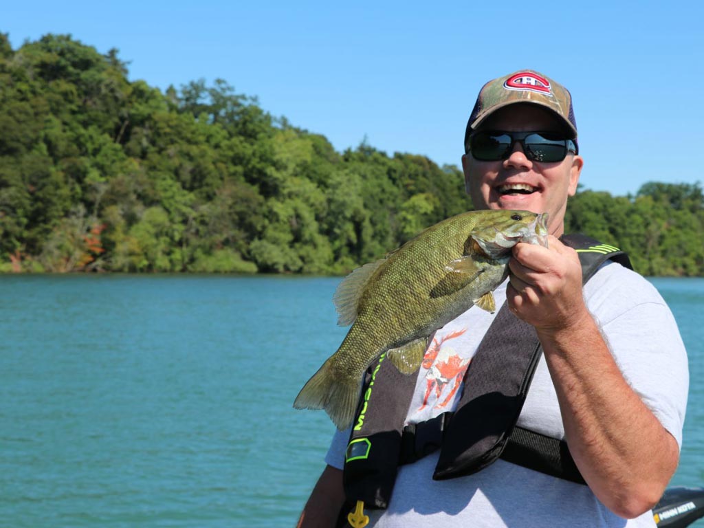 A man holding a Smallmouth Bass and smiling with beautiful azure water and a forest behind him.